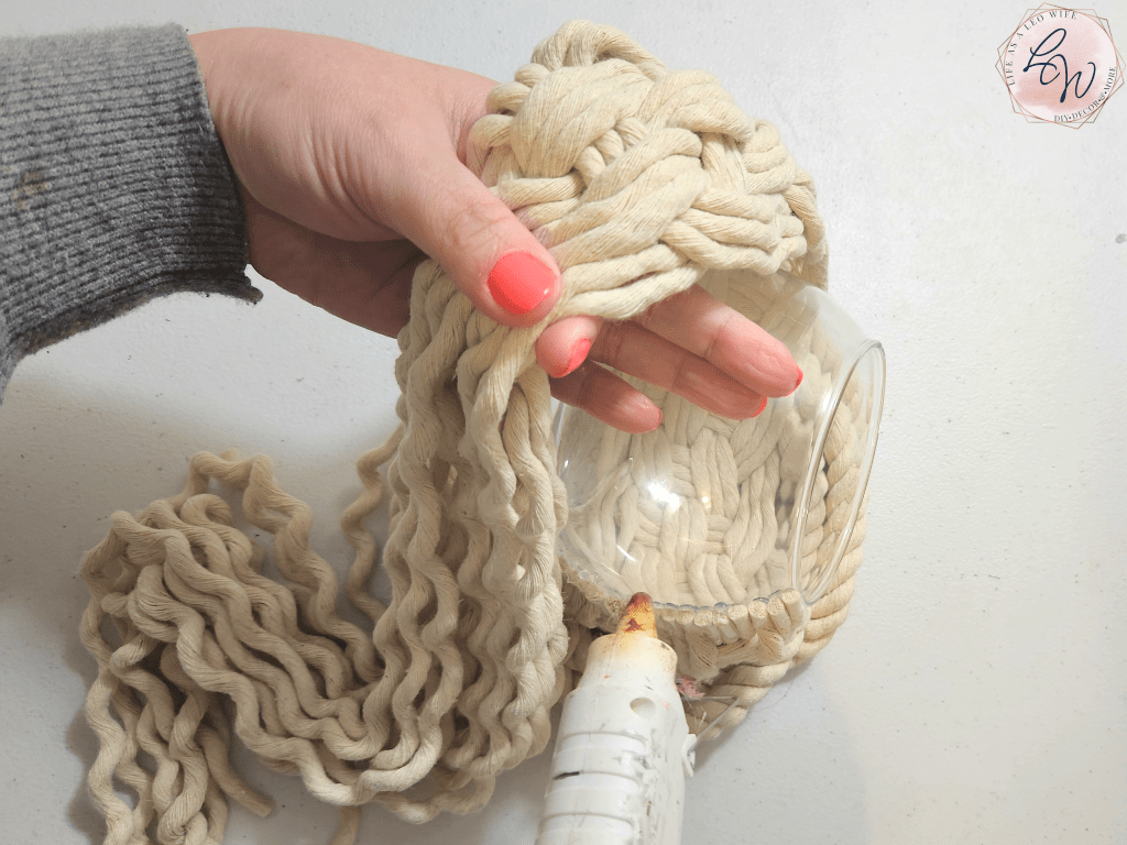 Adding hot glue to the vase while holding the finished knot patterned ropes out of the way before gluing them down.