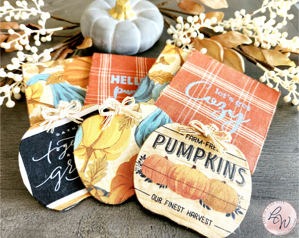 Fall coasters with a concrete pumpkin and fall stems behind them: 2 square coasters with pumpkins and gourds, 2 square coasters with rust and tan plaid base with "let's get cozy" on one and "hello pumpkin" on the other, 1 pumpkin coaster with "gather together and be grateful" on it, 1 pumpkin with pumpkins and gourds, and one pumpkin coaster with "farm fresh pumpkins" on it.