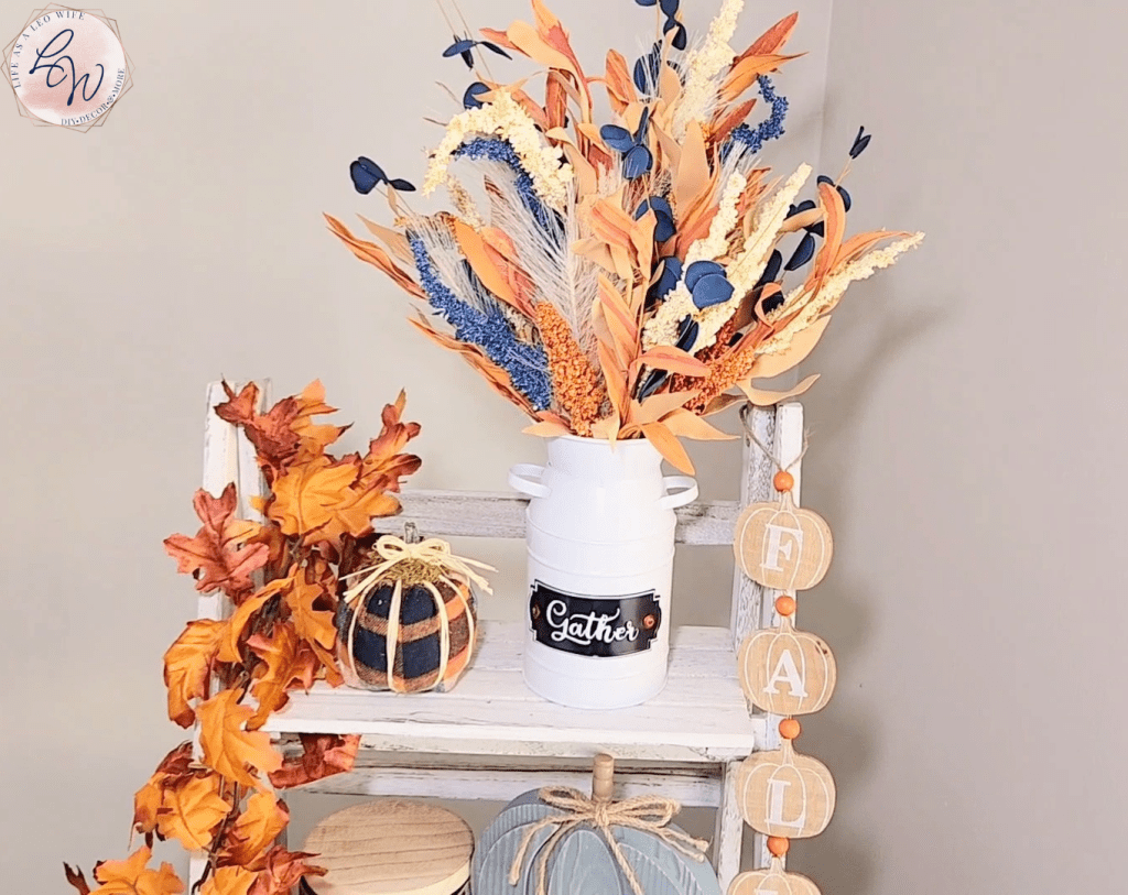 Top shelf of ladder shelf with a white milk jug vase with blue, rust, and coral stems in it sitting next to the finished flannel shirt sleeve pumpkin.