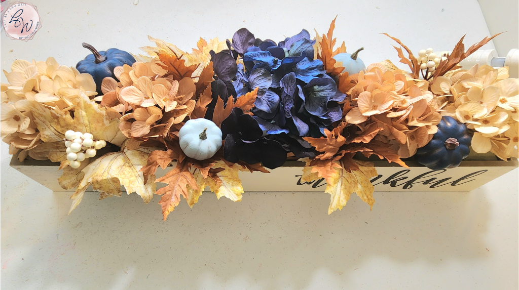 Fall wood planter box with "thankful" on the front filled with tan, copper, and navy hydrangeas, blue pumpkins, and leaf stems, sitting on a desk, viewed from above.