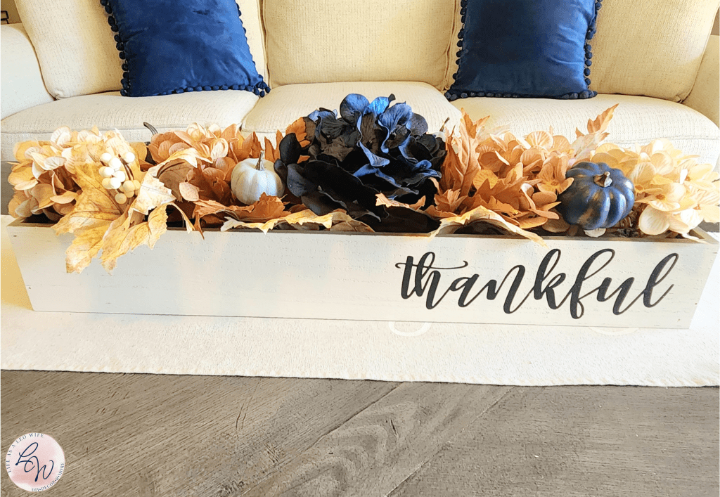 Fall wood planter box with "thankful" on the front filled with tan, copper, and navy hydrangeas, blue pumpkins, and leaf stems, sitting on a farmhouse style coffee table.