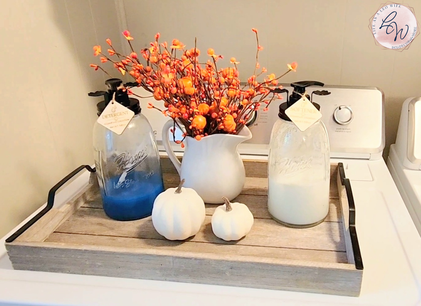 Laundry detergent and fabric softener in dispensers in decorative jars on a wood tray with pumpkin stems and white pumpkins in my fall laundry area during the home tour.