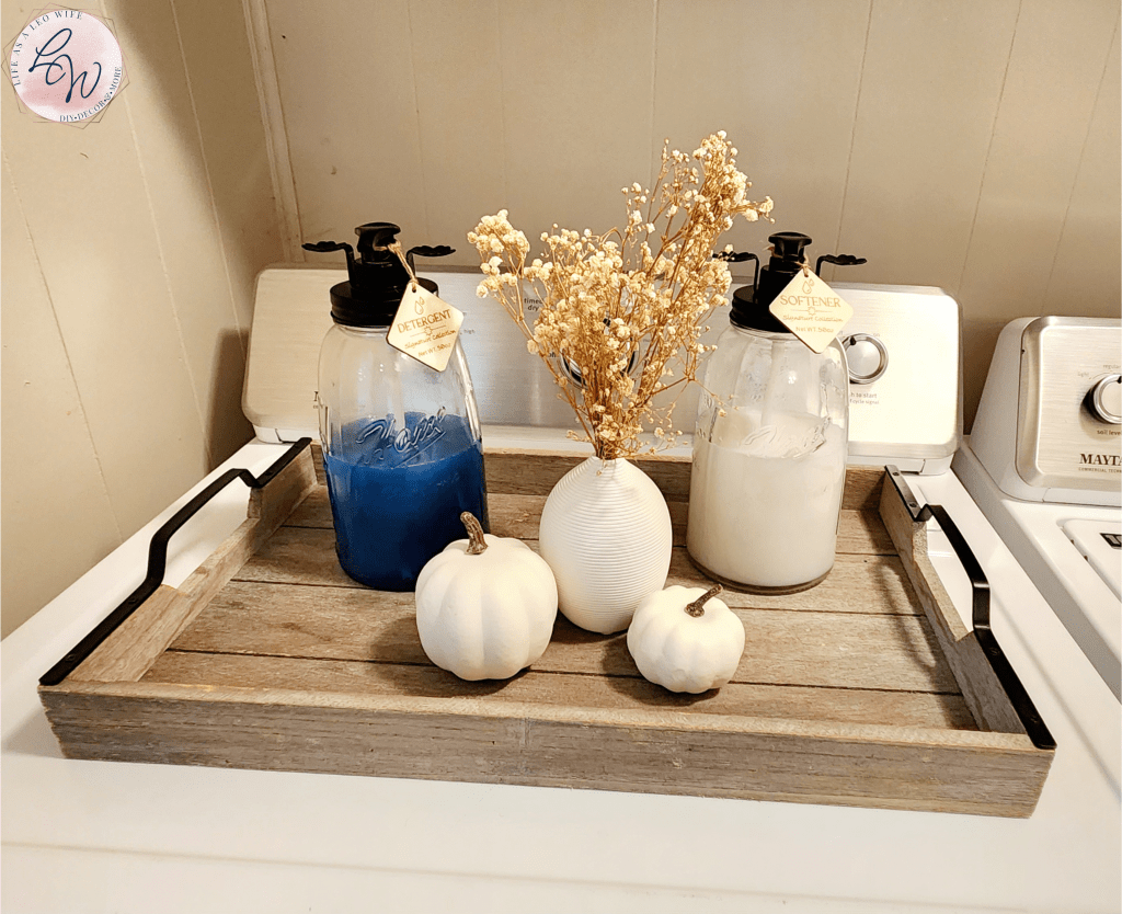 Tray on top of a dryer with large glass mason jars with dispensers and tags for laundry detergent and fabric softener with a vase & baby's breath and fall pumpkins on either side.