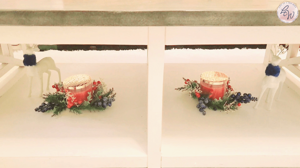 Bottom of coffee table with 2 sides & a Christmas candle sitting in a greenery candle ring with a white reindeer with a cobalt blue fur collar sitting next to it on each side.