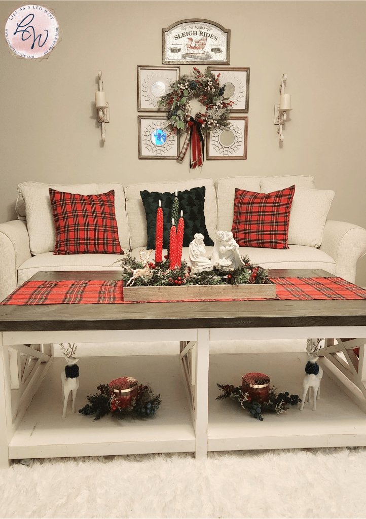 Christmas decor in a living room with plaid pillows & a hunter green pillow on a white couch, a decorative mirror set above it with a Christmas wreath with plaid & velvet bows hung from them & farmhouse candle sconces flanking them, & finally, a coffee table with a plaid runner under a wood tray filled with greenery, a nativity set, & an advent candleholder on top & candles with greenery rings and white reindeer in each shelf on the bottom.
