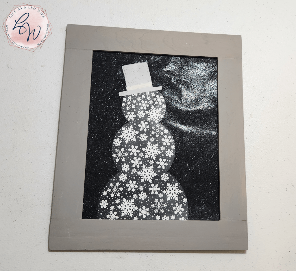 Christmas gift bag, with a black glitter background & a snowman made of snowflakes, turned into wall art with a gray wood frame.