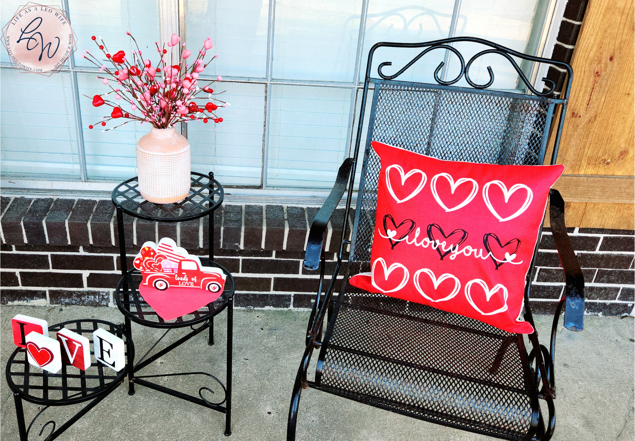 Valentine's Day porch chair with a red "love" pillow sitting in it & a 3-tier table to the left of it with a vase full of Valentine's heart branches on the top tier, a Valentine's truck on the 2nd, & l-o-v-e blocks on the bottom tier.