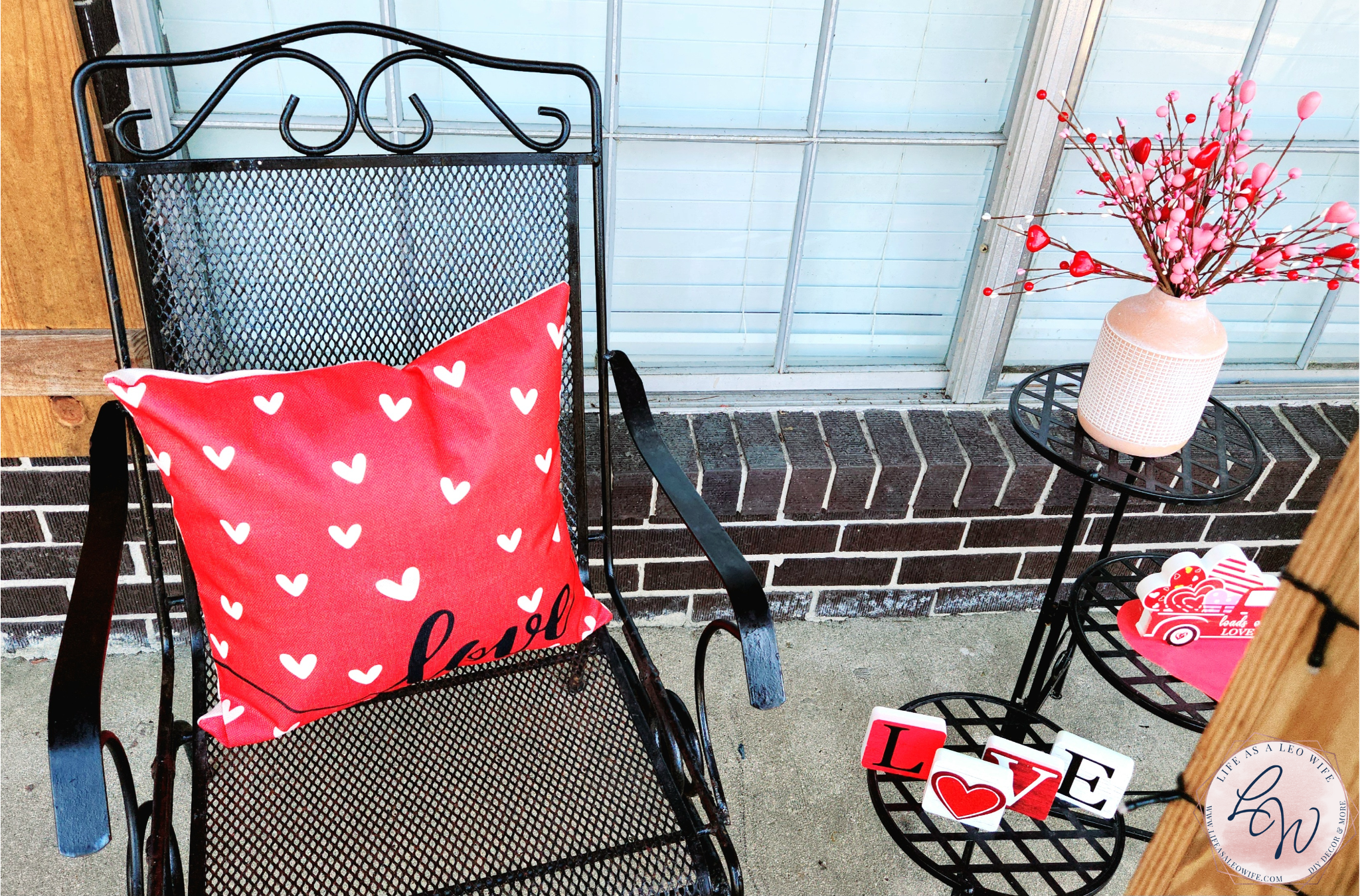 Valentine's Day porch chair with a red "love" pillow sitting in it & a 3-tier table to the right of it with a vase full of Valentine's heart branches on the top tier, a Valentine's truck on the 2nd, & l-o-v-e blocks on the bottom tier.