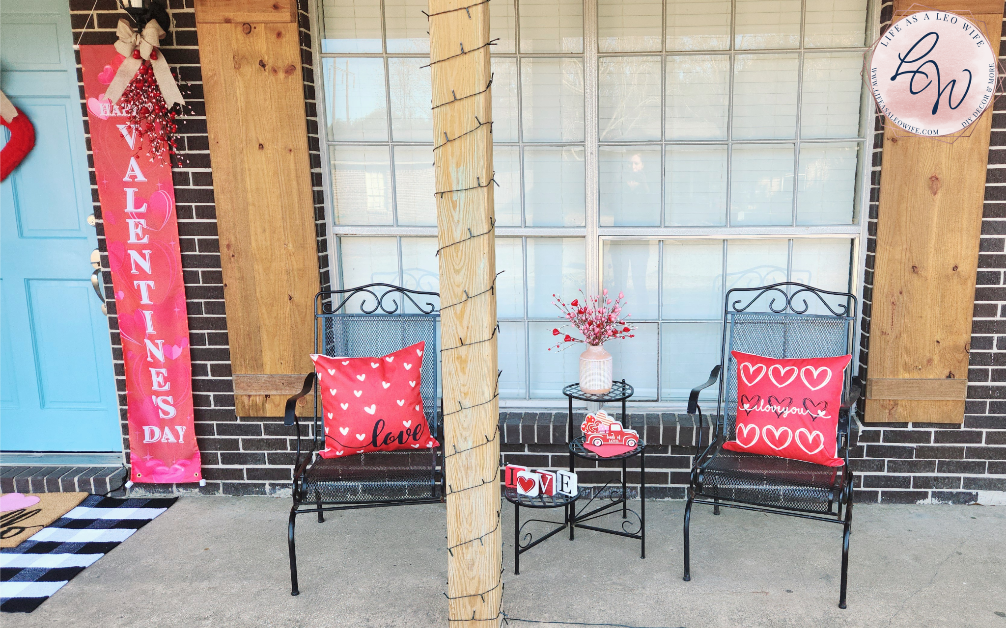 Part of the front door & one V-Day banner, and V-Day porch chairs with red "love" pillows sitting in them & a 3-tier table next to it with a vase full of Valentine's heart branches on the top tier, a Valentine's truck on the 2nd, & l-o-v-e blocks on the bottom tier.