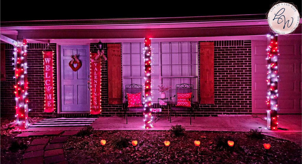 Valentine's Day porch at night with red & white lights wrapped around 3 columns, banners hanging on either side of the door with lights & "welcome" on 1 & "Happy Valentine's Day" on the other, a red heart wreath on the door, a black & white buffalo check rug in front of the door, & a teardrop Valentine's swag hanging from the porch light, 2 porch chairs in front of large windows with red pillows with "love" & white hearts & a table between the chairs with a vase containing Valentine's branches of red & pink hearts on the top tier, a Valentine's farmhouse truck on the 2nd, & "l, o, v, & e" letters on the bottom tier, & finally a set of heart solar lights running in front of the porch.