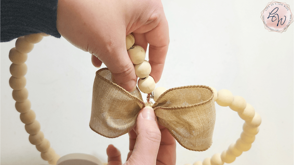 Attaching a khaki bow with a pipe cleaner to the metal clasp that attaches the two sides of the wood bead wreath that I formed into a heart.