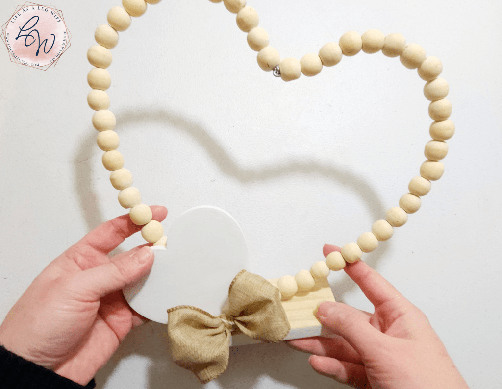 Wood bead heart attached to a wood base with a wood heart attached to the left side of the wood bead heart and the base and a khaki linen bow glued over the part where the wood heart and the base meet.