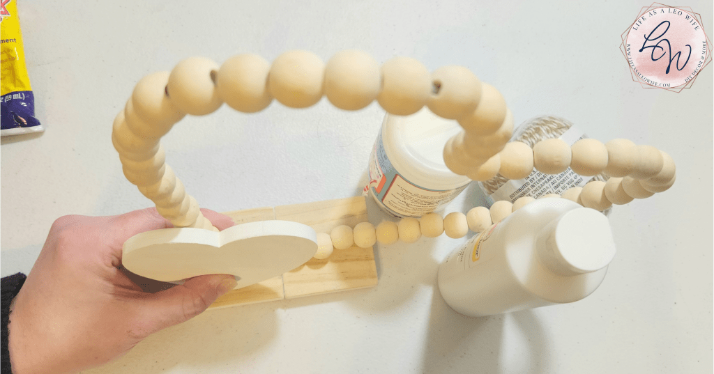 Pressing a white wood heart onto a wood bead wreath bent into a heart shape and attached to the wood base to hold up the shelf sitter decoration.