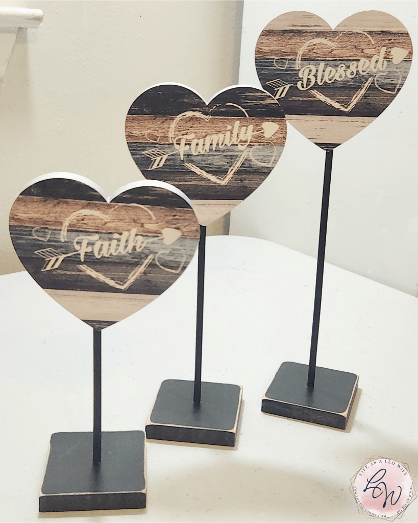 Three wood heart topiary-like decorations; wood hearts with brown, khaki, dark gray, and white painted horizontal stripes with family, faith, and blessed written on each one. Each heart is on a wood stick that is inserted into the bottom of it and into the top of a wood base.