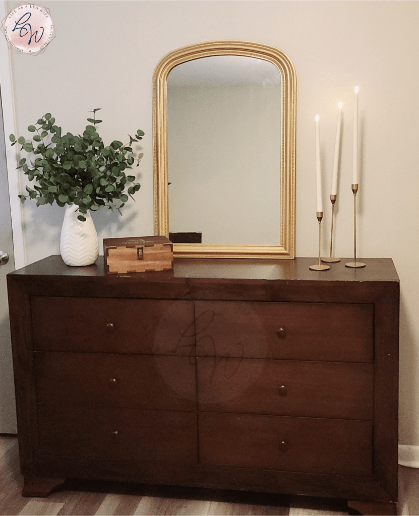 Six-drawer dresser with a vase full of eucalyptus, a wood box, gold mirror, & 3 gold candlesticks with ivory faux candles on top.