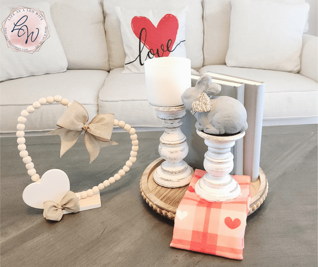 Faux concrete bunny with a twine heart around its neck sitting on a farmhouse candleholder which is on a Valentine's Day tea towel with other decor on a tray and sitting next to a wood bead heart decoration in a Valentine's vignette.