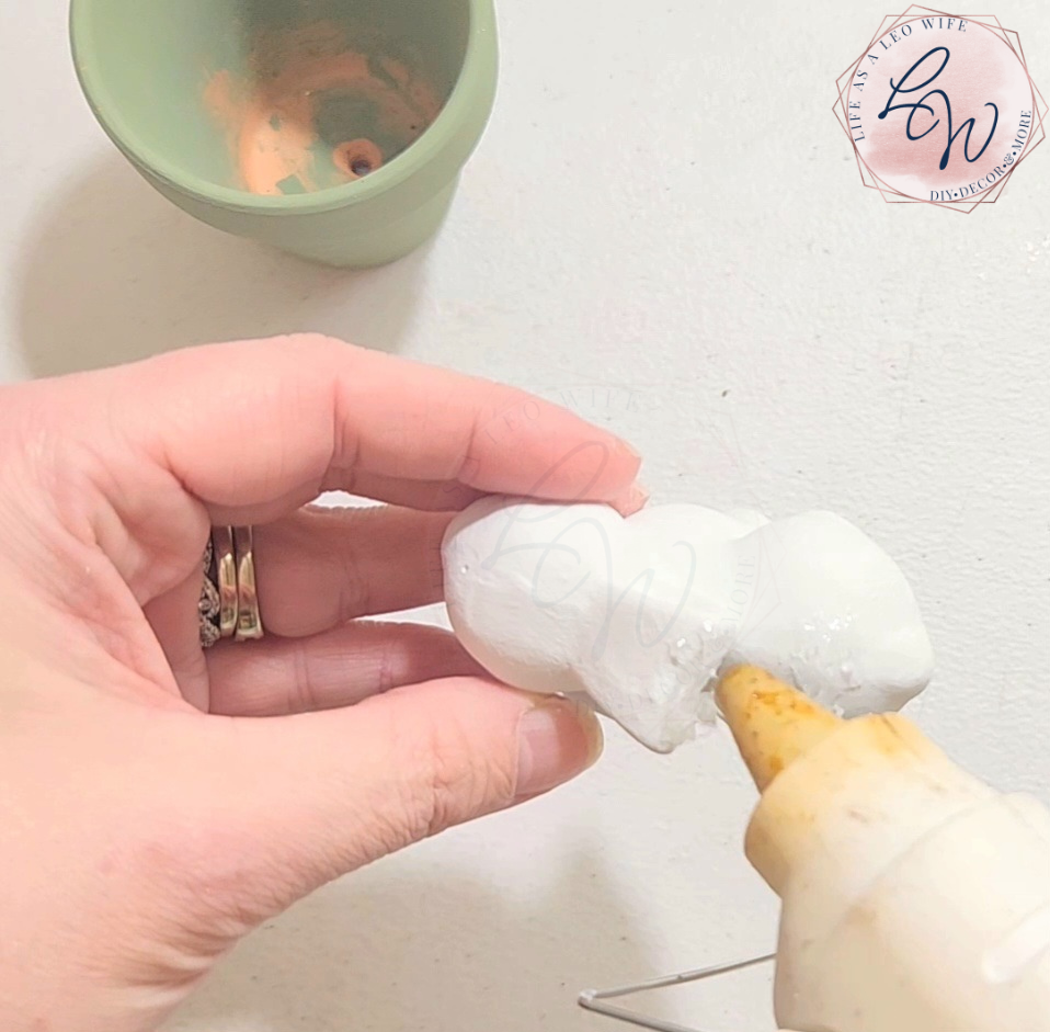 Adding hot glue on top of a white ceramic bunny to place a small Dollar Tree terracotta pot on top to create a DIY bunny flowerpot.