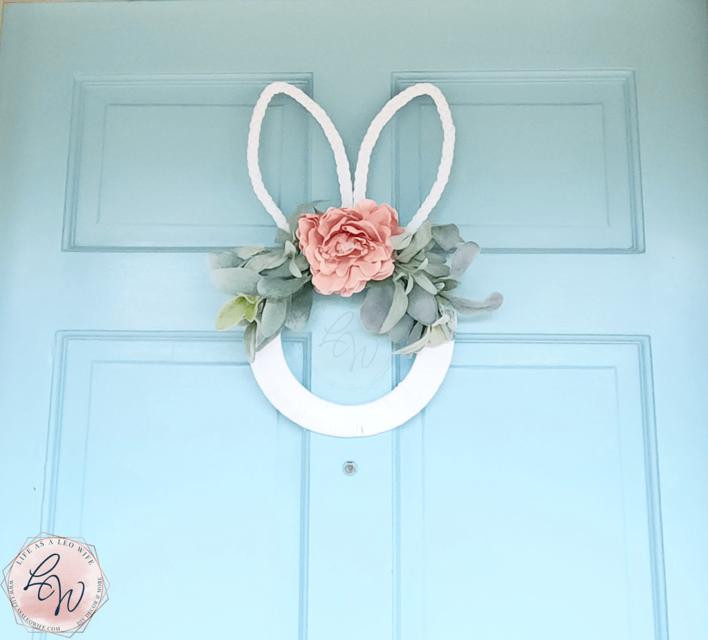 Finished bunny wreath hanging on the front door. The wreath is wrapped in white macrame cord around the center ring with braided macrame cord covering the bunny ears at the top & lamb's ear greenery extending out of the middle just below the ears with a dusty pink dahlia flower in the center of the greenery,