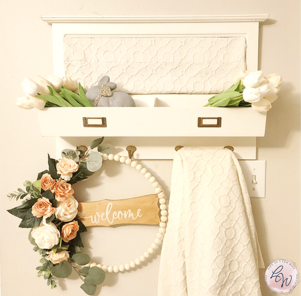 Easter & spring entryway decor- white wood shelf with tulips in the shelves and wood bead wreath with "Welcome" sign and roses on it and cream fabric hanging from hooks.