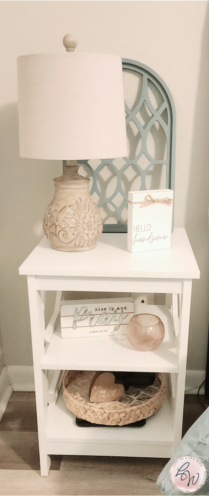 Nightstand on the left side of the master bed with a wood arch piece behind a tan lamp with flowers on the base, and other farmhouse decor signs on all 3 shelves.