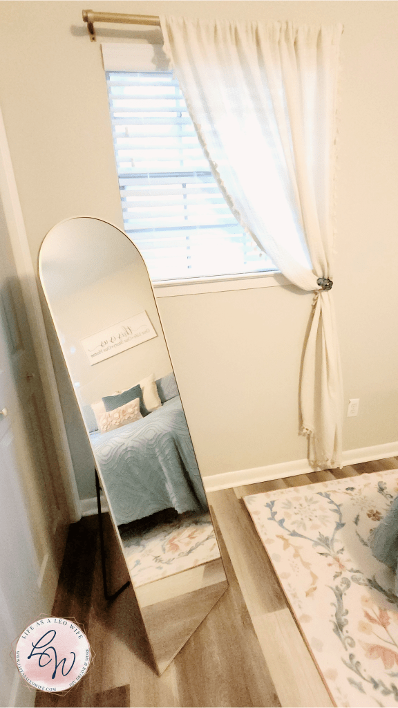 Left side of the master bedroom refresh reveal with a gold full-length mirror in front of a window with farmhouse curtains pulled to the side opposite the mirror and tied back with a flower tieback.
